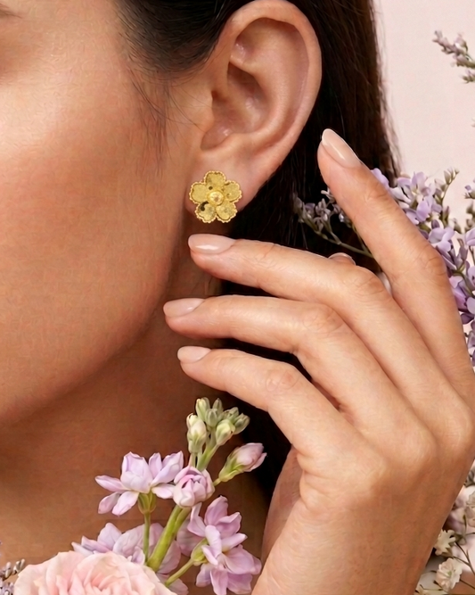A close-up lifestyle shot of a woman wearing a textured gold flower-shaped stud earring, surrounded by soft pink roses and delicate purple and white flowers against a pale pink background.